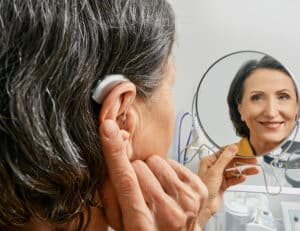 Mature woman with BTE hearing aid looks at herself in mirror and tries on hearing device, close-up. Hearing loss treatment
